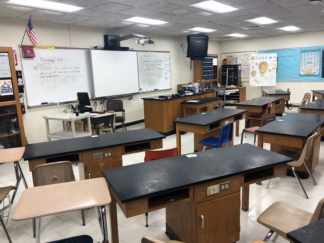 A high school classroom is filled with educational posters, a long white board and desks with electrical outlets.