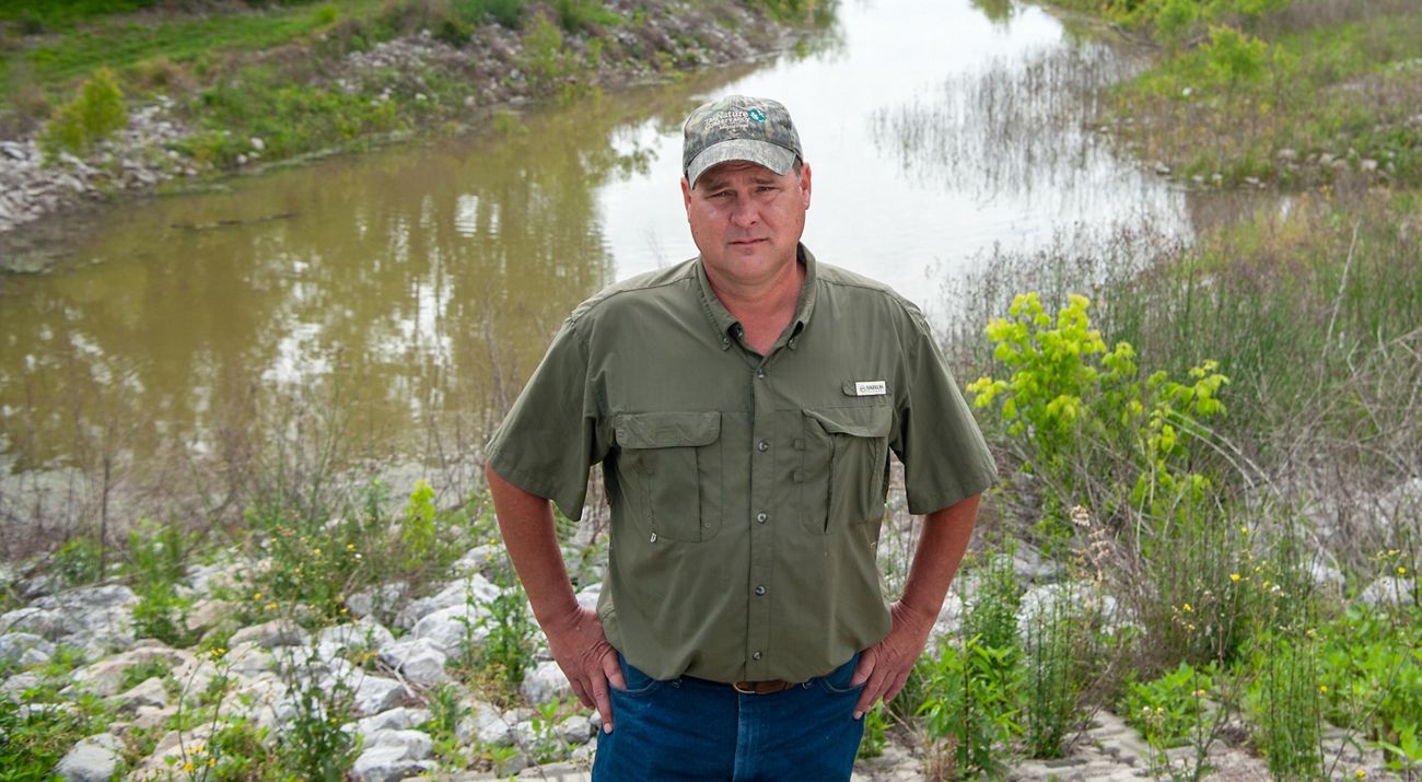 A man wearing a hat and a green shirt stands in front of a wetland.