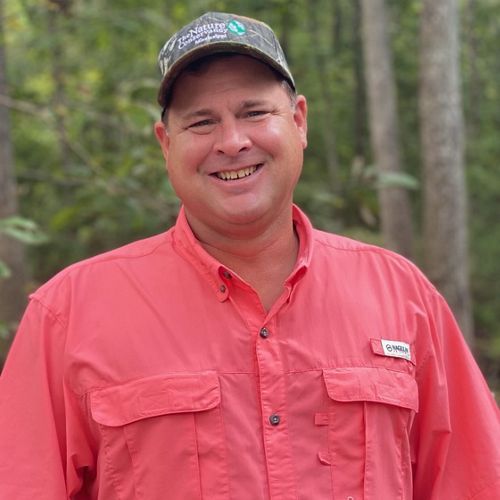 A man wearing a salmon colored shirt and a cap smiles at the camera.