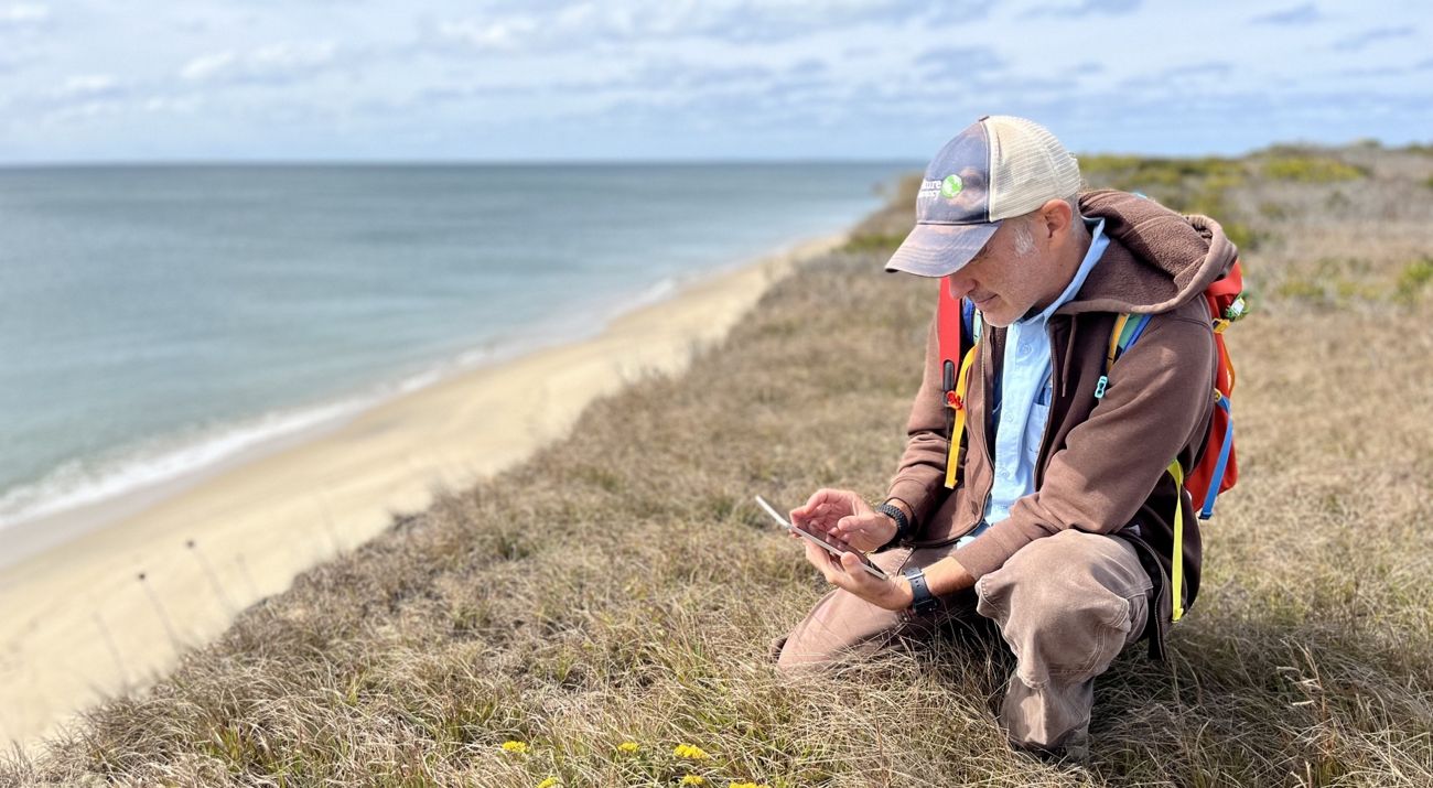 Rich Couse, seasonal stewardship staff member, kneels in the grass of a dune along the beach on Tuckernuck Island on a sunny day.