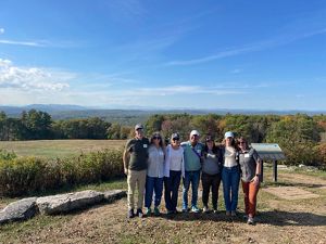 Seven adults pose in a line on a sunny day with a green landscape behind them.