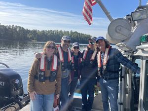 Five adults wearing life jackets pose together on a boat on a sunny day.
