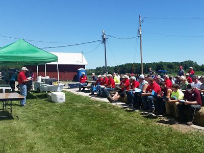 Man speaking to large group of farmers in front of farm field.