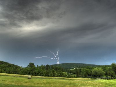 Lightning forks from a heavy gray sky arcing behind a green mountain ridge. A line of trees runs between two open fields.