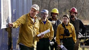 A man in yellow fire gear points to a map hanging on a wall during a pre-burn briefing. Four people stand behind him listening to the briefing.