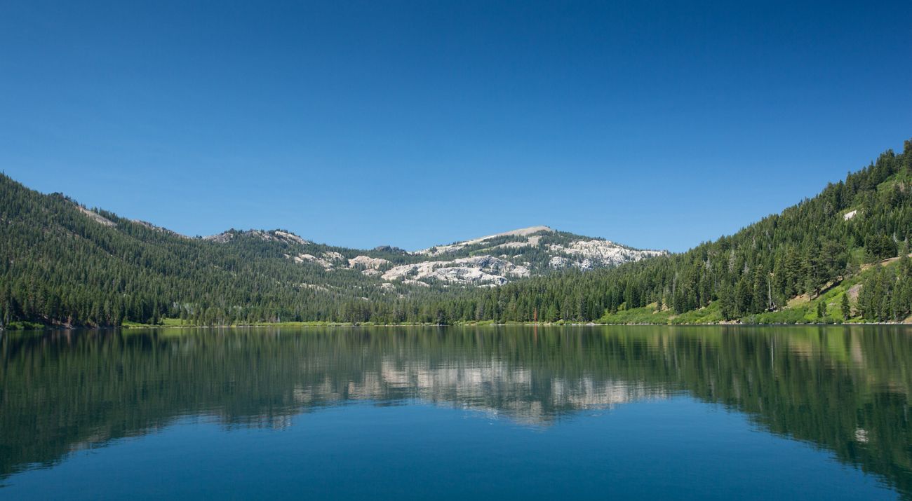 A tranquil lake surrounded by forest-covered mountain slopes under clear blue skies.