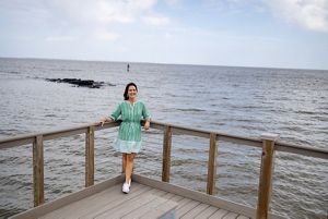 Jennifer VanVrancken, in a striped green dress and white tennis shoes, stands on the corner of a boardwalk over the water. In the background, a pile of rocks pokes above the rippling water. 