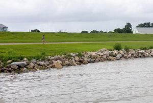 A shot of the Metairie waterfront. Rocks are piled on the shore to protect against waves and storms. A jogger runs down a pathway in front the tall grassy levee. 
