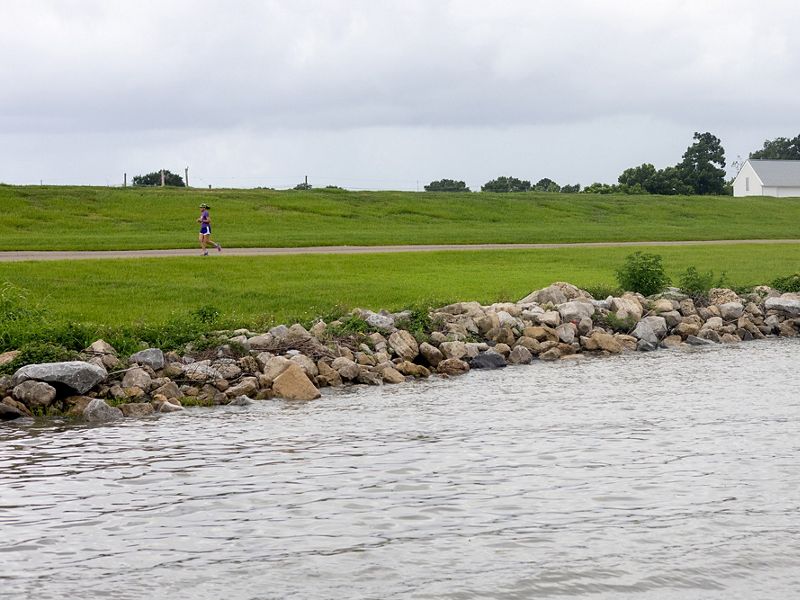 A shot of the Metairie waterfront. Rocks are piled on the shore to protect against waves and storms. A jogger runs down a pathway in front the tall grassy levee.