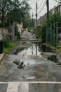 Large puddles carpet a wide, paved alleyway with houses on either side. 