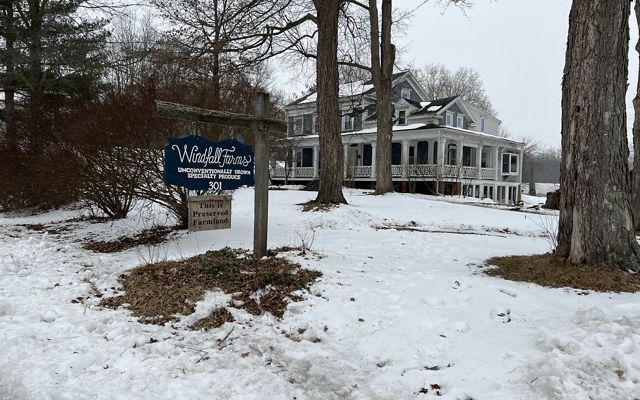 Snow covers the foreground of a lawn and walkway leading to a gray and white farmhouse seen in background. A sign at center says 'Windfall Farms: Unconventionally Grown Specialty Produce.'