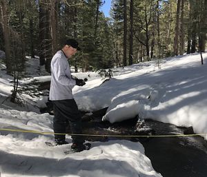 A man wearing a black hat stands in snow next to a small creek.