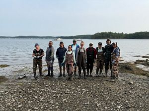 A group of ten people in waders stand on the shore next to a body of water.