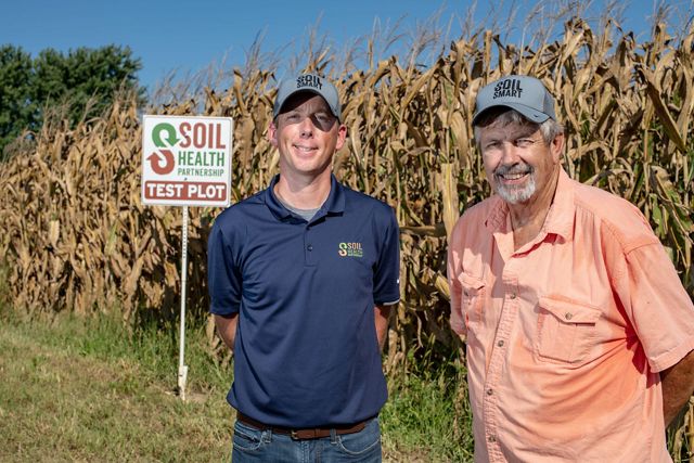 Two men with hats stand beside rows of corn, a sign near them for the Soil Health Partnership test plot.