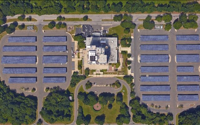 Aerial view of a 5 rows of solar arrays surrounded by trees with a building in the center.
