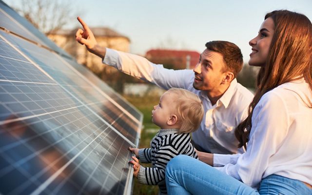 Man shows his family the solar panels on the plot near the house during a warm day. Young woman with a kid and a man in the sun rays look at the solar panels. 