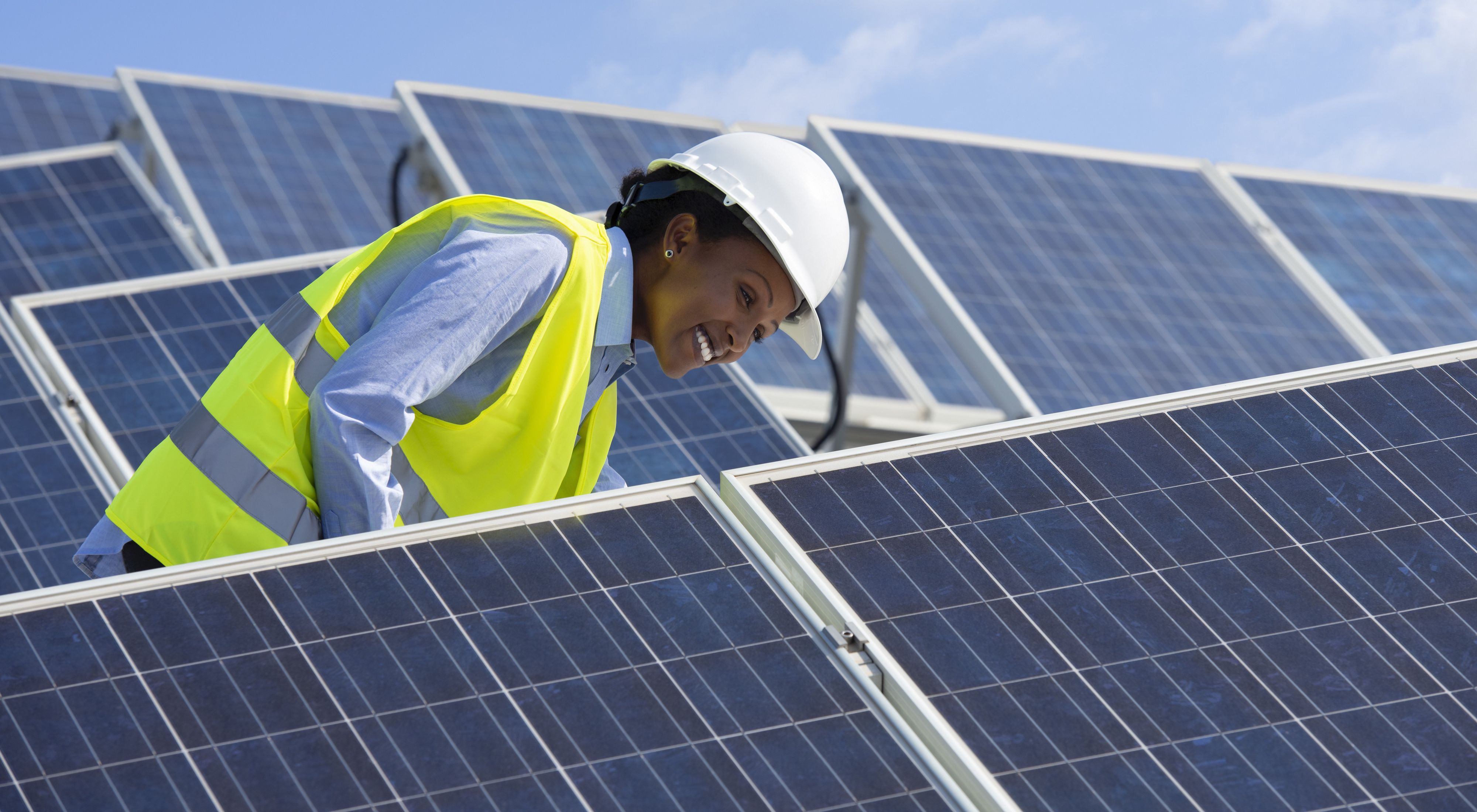 Woman with hard hat smiles while standing amongst solar panels. 
