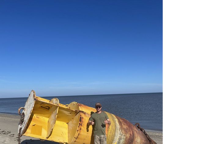 A photo of Russel on the beach in front of a large metal object that has washed up to shore.