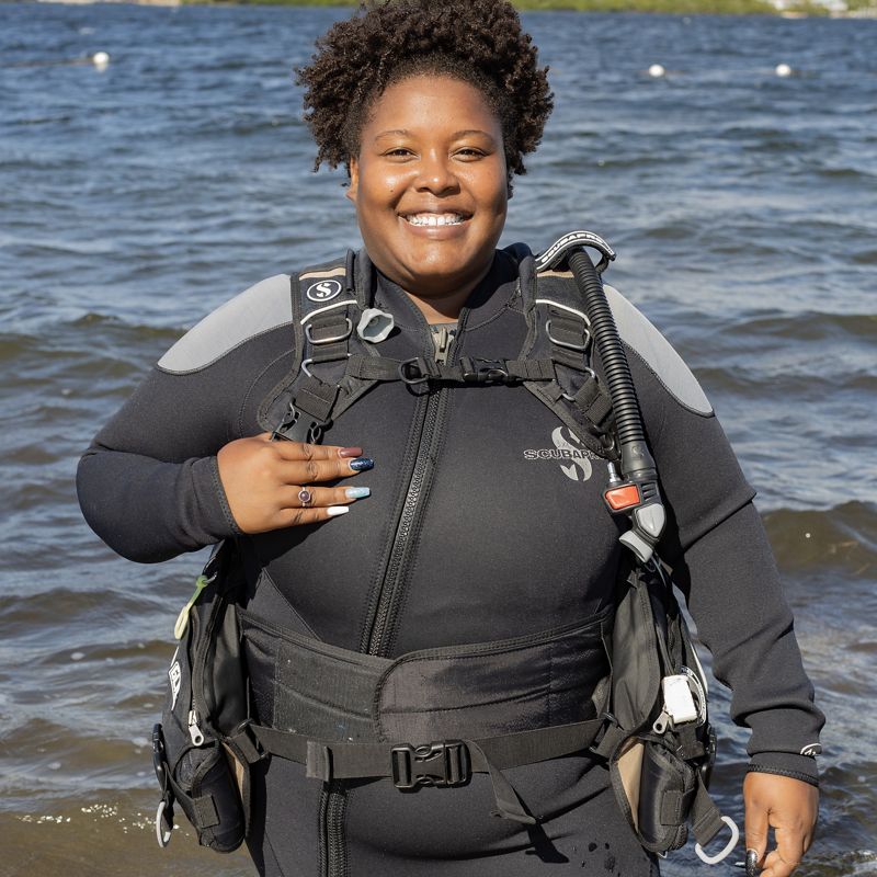 Tiara posing for a photo wearing a wet suit and scuba gear with the ocean behind her.