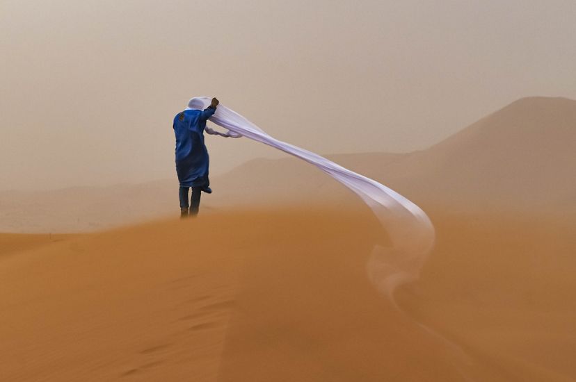 a person in blue seen from behind on amber sand, with white head scarf unraveling in the wind