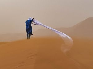 a person clothed in blue walks through an amber desert sandstorm with a white scarf blowing behind