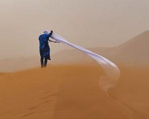 Una persona vestida de azul camina a través de una tormenta de arena del desierto con un pañuelo blanco soplando detrás.
