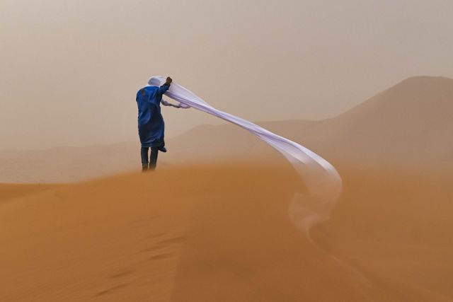 a person in blue seen from behind on amber sand, with white head scarf unraveling in the wind