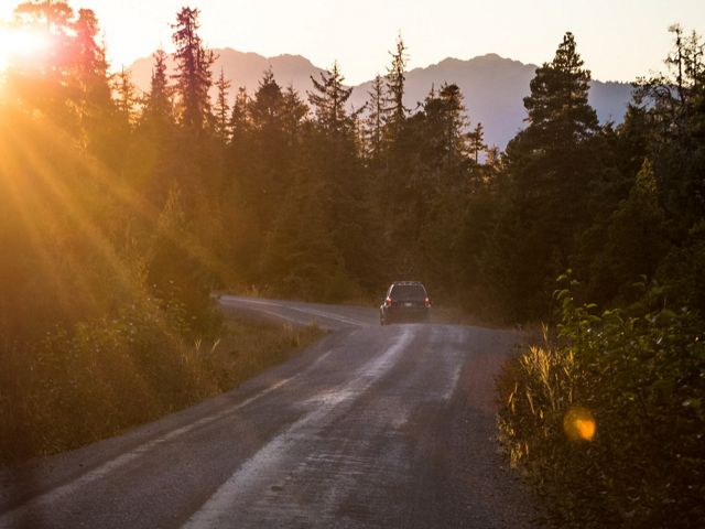 A car drives down a dirt road that cuts through a forest as the sun sets over the tree line.