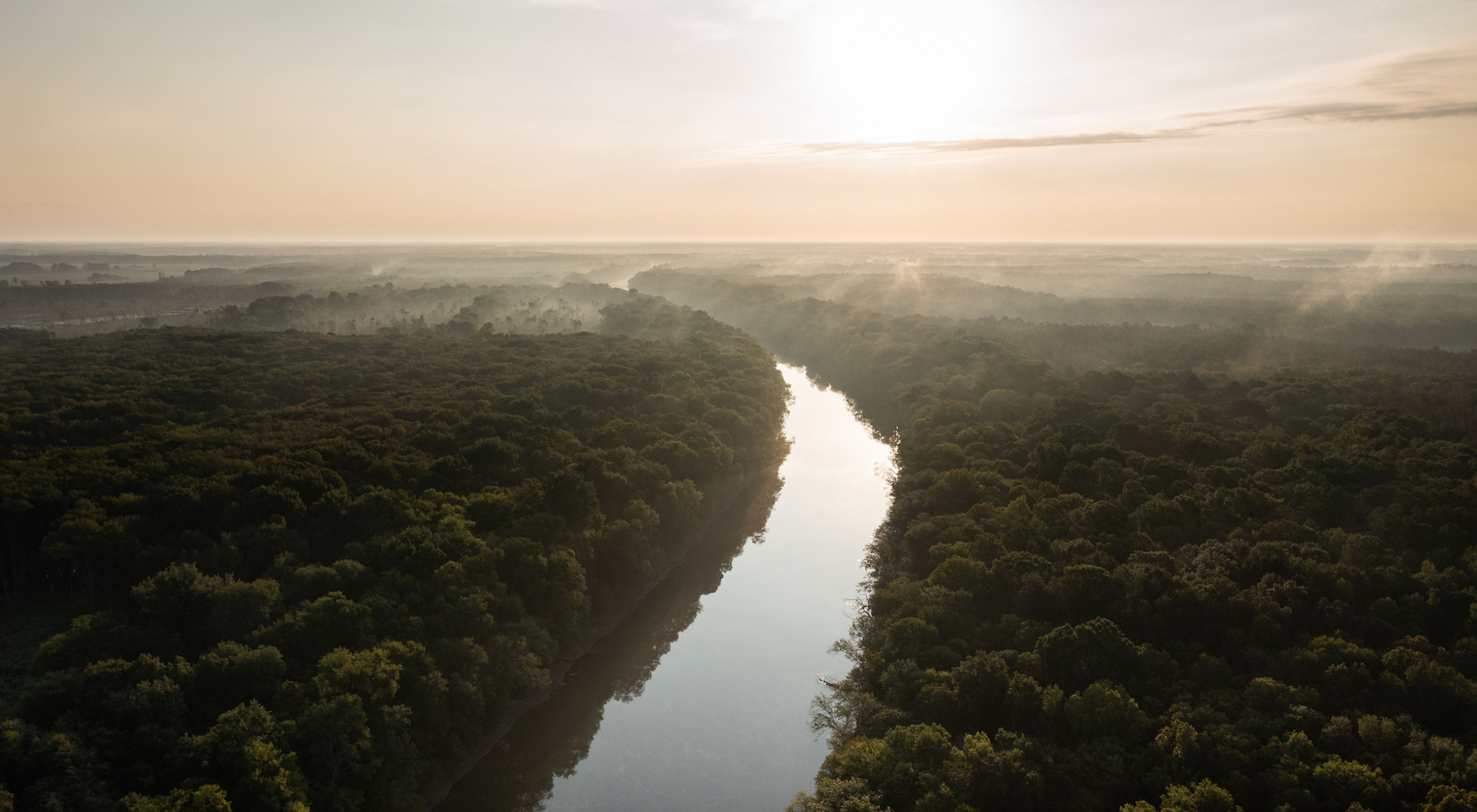Aerial view of a river flowing through a forest while mist rises off the trees under a hazy orange sky.