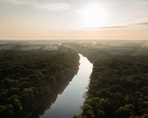 A wide flat river gently curves through a forest. Morning mist rises above the trees.
