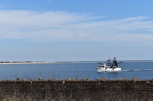 A brick wall borders blue waters; a large white shrimp boat floats in calm waters.