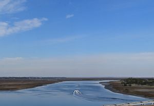 Brown marsh grasses border the blue waters of a river as it meets the coast. Boats travel the river, leaving a white wake behind them.