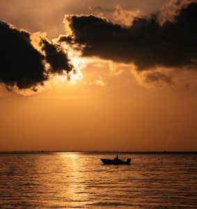 The black silhouette of a boat travels slowly on calm waters, reflecting the bright orange sunset.