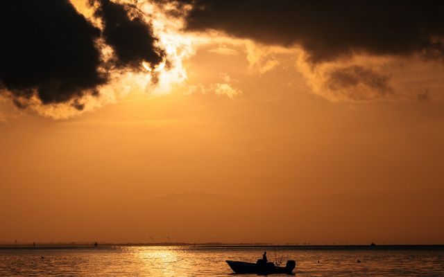 The black silhouette of a boat travels slowly on calm waters, reflecting the bright orange sunset.