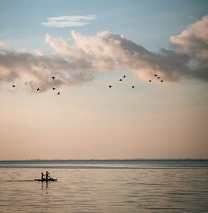 Two black silhouettes of people sit on a paddleboard, above them are thirteen black birds flying in a cloudy evening sky.