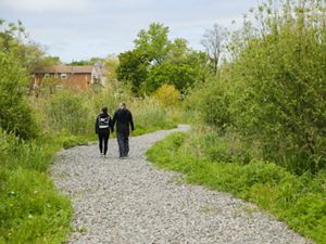Couple, holding hands, walks on a gravel path surrounded by greenery, with a brick house in the background.
