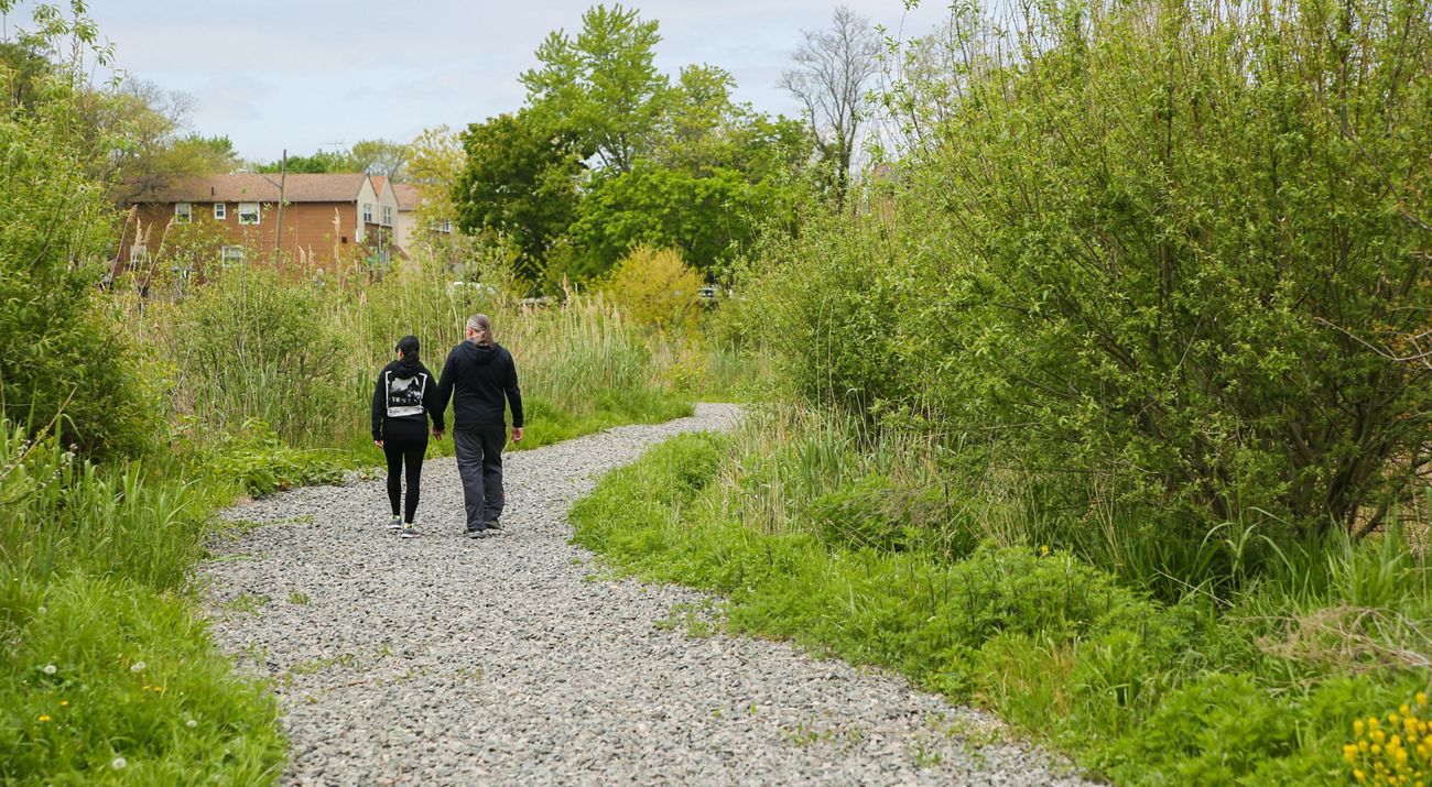 Couple, holding hands, walks on a gravel path surrounded by greenery, with a brick house in the background.