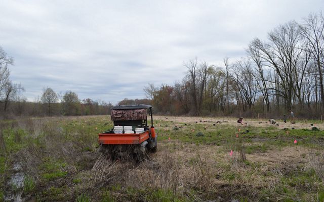 A small utility vehicle carrying buckets sits in a sparse field with people planting trees in the background.