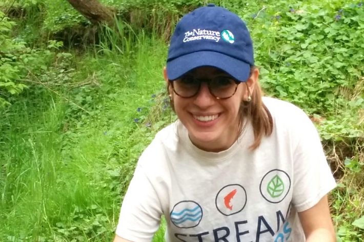 A smiling woman crouches down at the edge of a stream. She is holding one end of a white tube used to sample water quality.