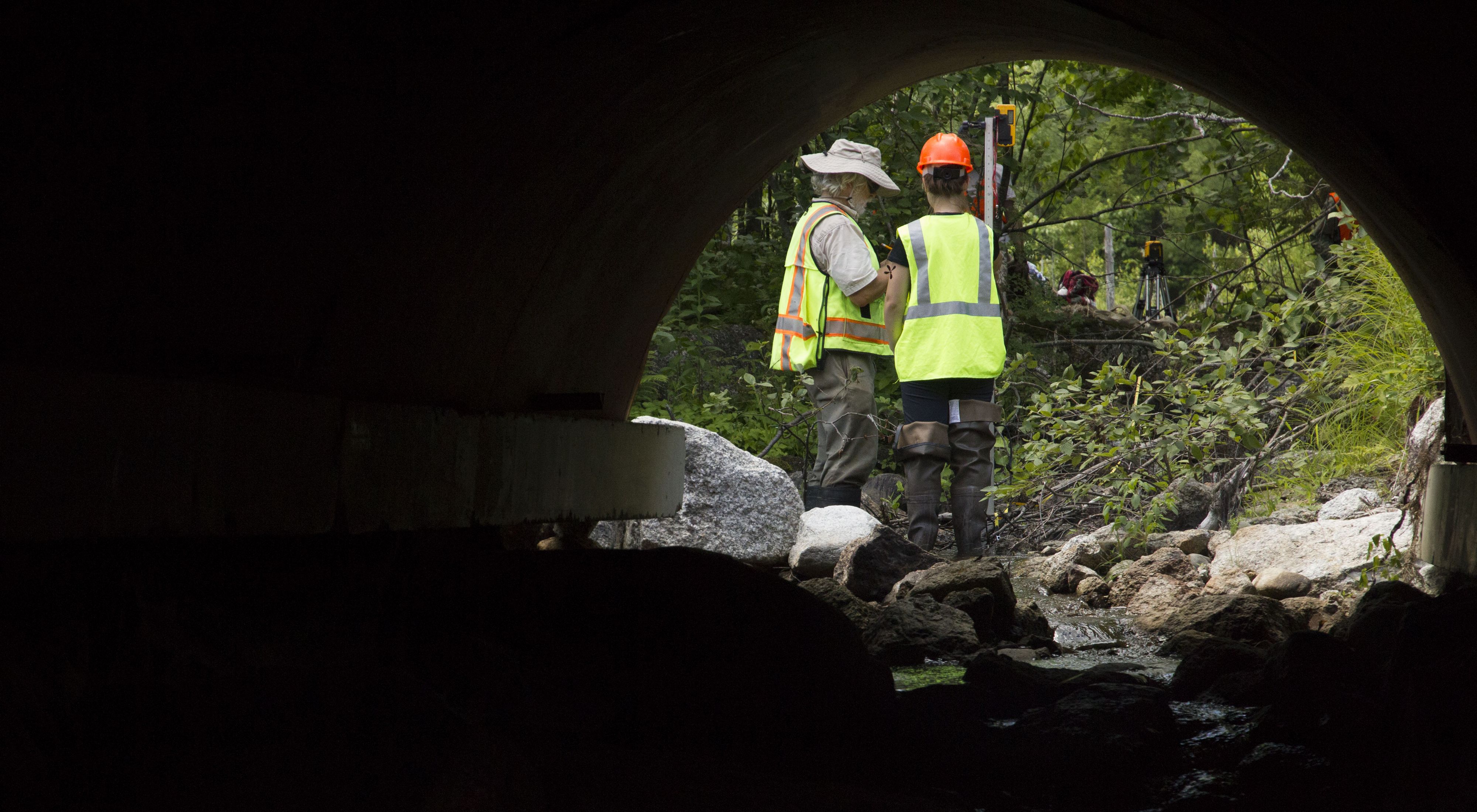 Two construction workers viewed from inside a tunnel under a road.