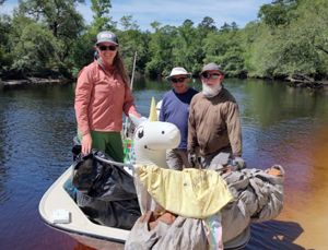 two men and one women standing up on a boat with a river and threes in the background.