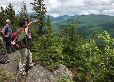 Three people stand on a rock outcrop looking out over a valley and rolling green mountain ridges. One person points ahead into the distance.