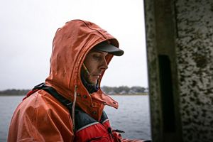 Steve Kirk wears an orange rain jacket, red life vest and black baseball cap and looks down. A coastal horizon line is in the background.