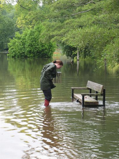 A person in boots wades through shin-deep floodwater past a partially submerged park bench.