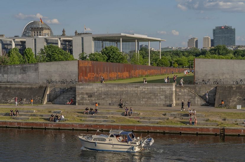 a river stretches in front of a concrete wall with bicycles and people milling about on the wall and in a field in the distance. Buildings and trees line the  horizon