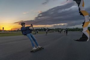 a person stands and rides a skateboard on a runway while holding onto a kite, sun sets in the background