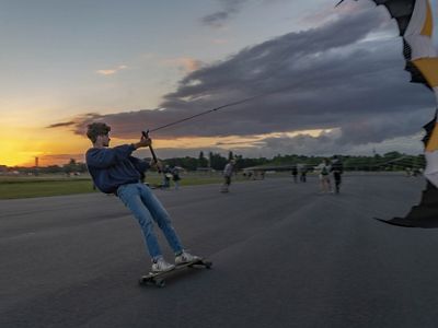 a person stands and rides a skateboard on a runway while holding onto a kite, sun sets in the background