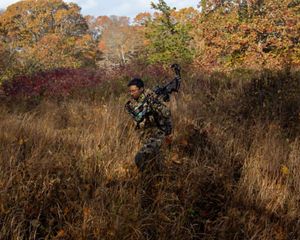man in camo walks through grassy field holding compound bow.