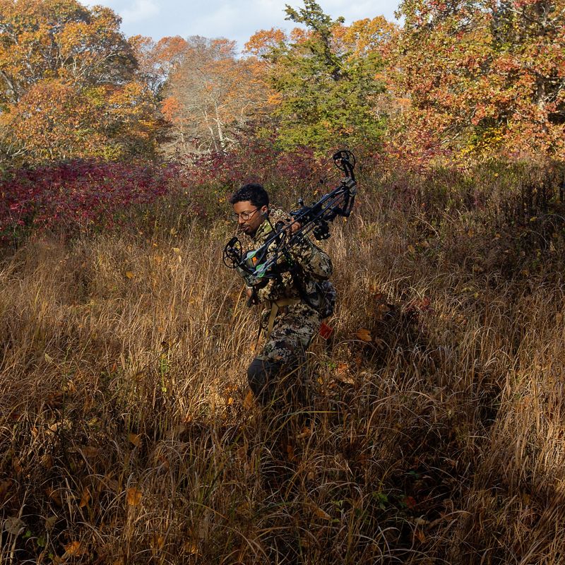 man in camo walks through grassy field holding compound bow.