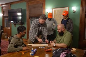 5 men gather around a table in a green room as one holds a small piece of metal in his hand.